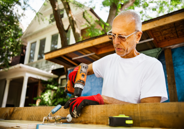 Suburban yard, older man with drill at wooden board, house and trees behind, midday sun, documentary style, people present.