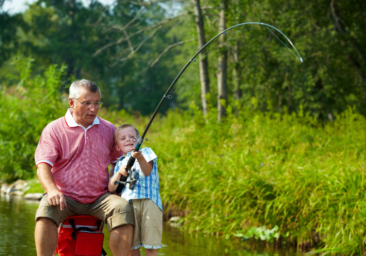 Riverside greenery, boy fishing with bent rod, man observes nearby, afternoon light, editorial travel photo, two people.