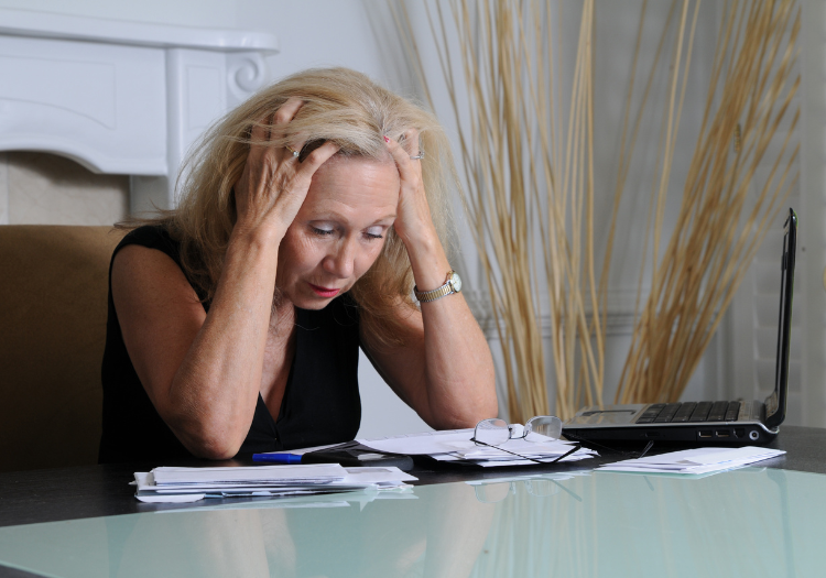 Office space, woman with head in hands, cluttered desk and laptop foreground, daytime lighting, documentary style, one person.