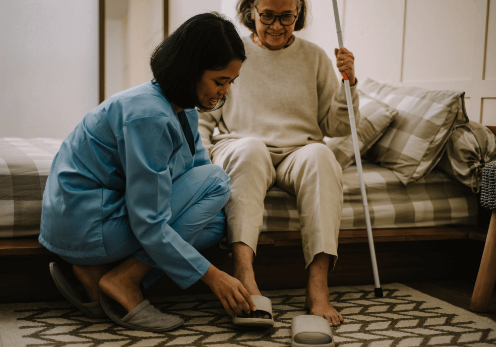 woman putting on slippers of an elderly woman walking away from elderly parent