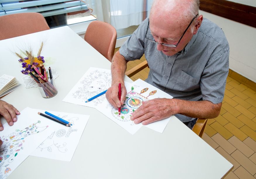 White table, elderly man coloring with pencils, drawings spread out, soft daylight, documentary style, one person.