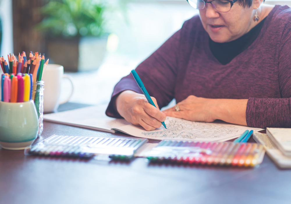 Kitchen table, older woman coloring, art supplies scattered, daytime natural light, documentary style, one person.