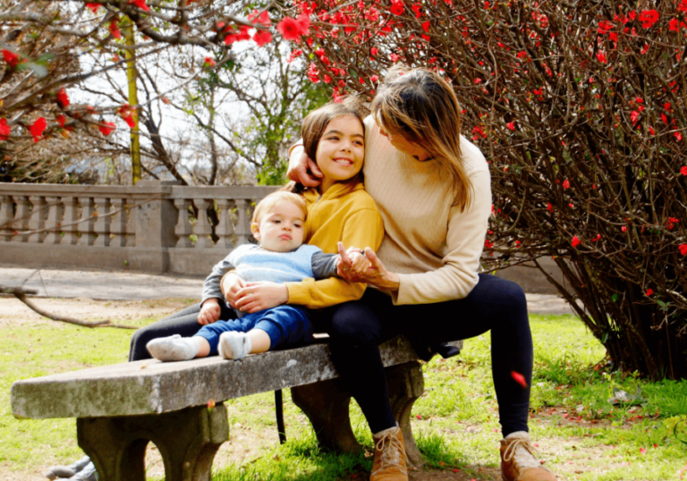 Garden bench, woman with baby, older child nearby, dappled sunlight, editorial travel photo, people present.