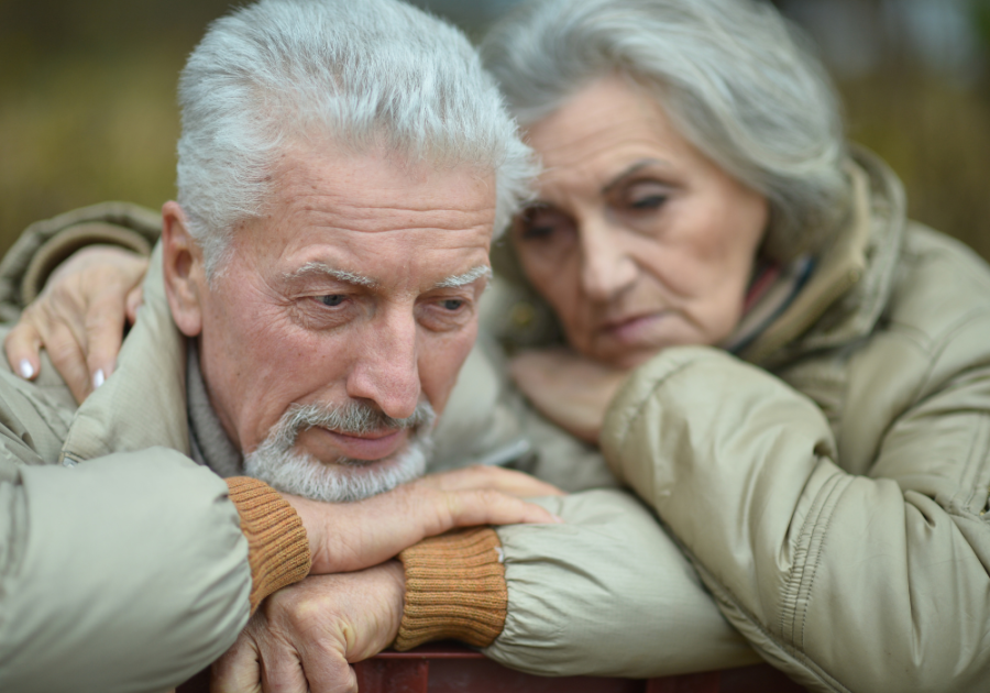 Park bench, elderly couple together, woman comforts man, overcast afternoon light, documentary style, people present.