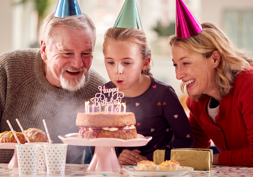 Birthday party table, girl blowing cake candles centered, warm indoor light, documentary style, two adults celebrating.