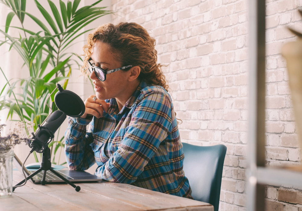 Bright room, wooden table, woman with curly hair podcasting, daytime natural light, documentary style, one person.