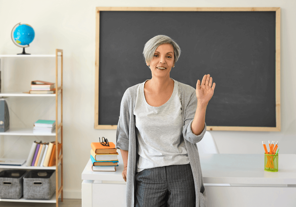 Classroom, older woman waving, desk with books foreground, soft morning light, documentary style photo, one person.