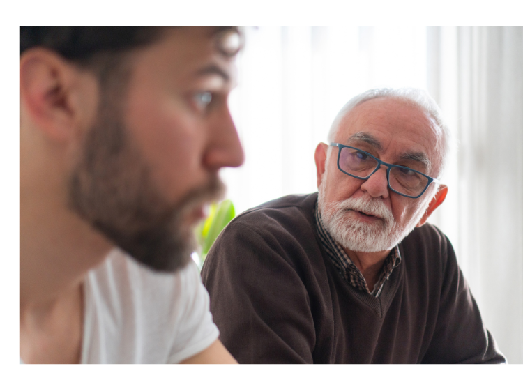 Well-lit room, two men in conversation, older man facing younger, daytime brightness, documentary style, people present.