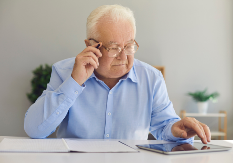 Home office, elderly man with glasses, seated at desk with tablet and papers, daylight, documentary style, single person.