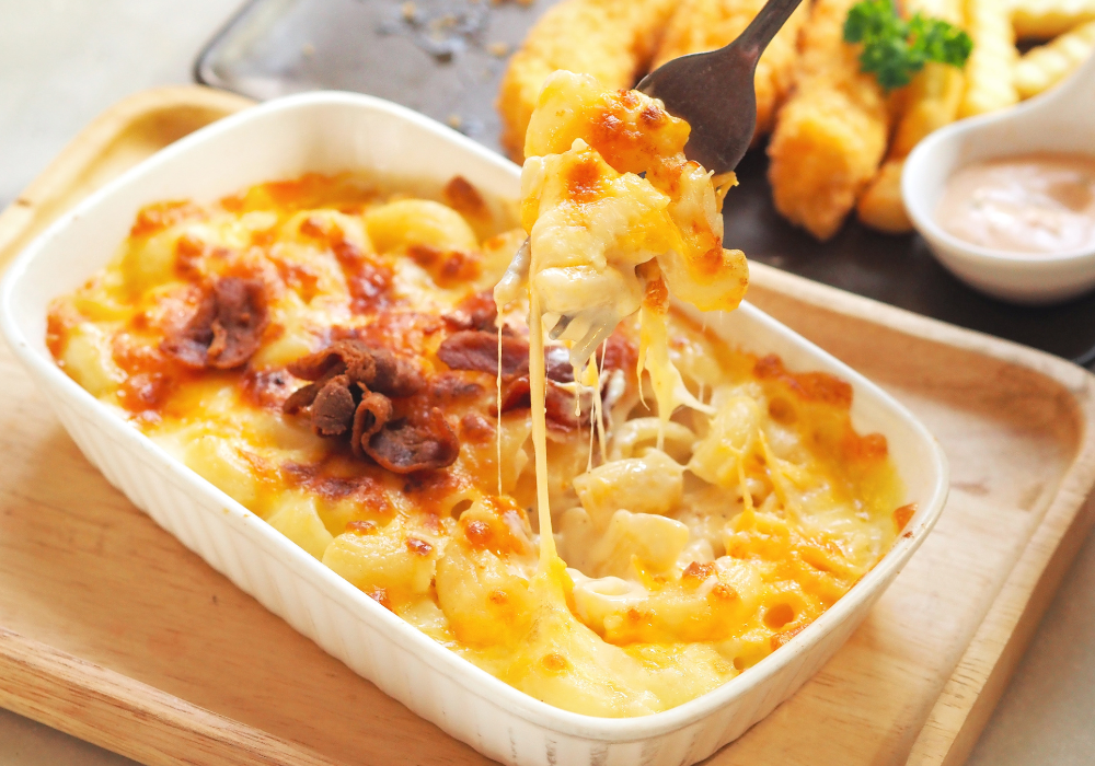 Restaurant table, cheesy bacon macaroni and cheese lifted by fork, white dish foreground with fries behind, bright midday light, editorial travel photo, no people.