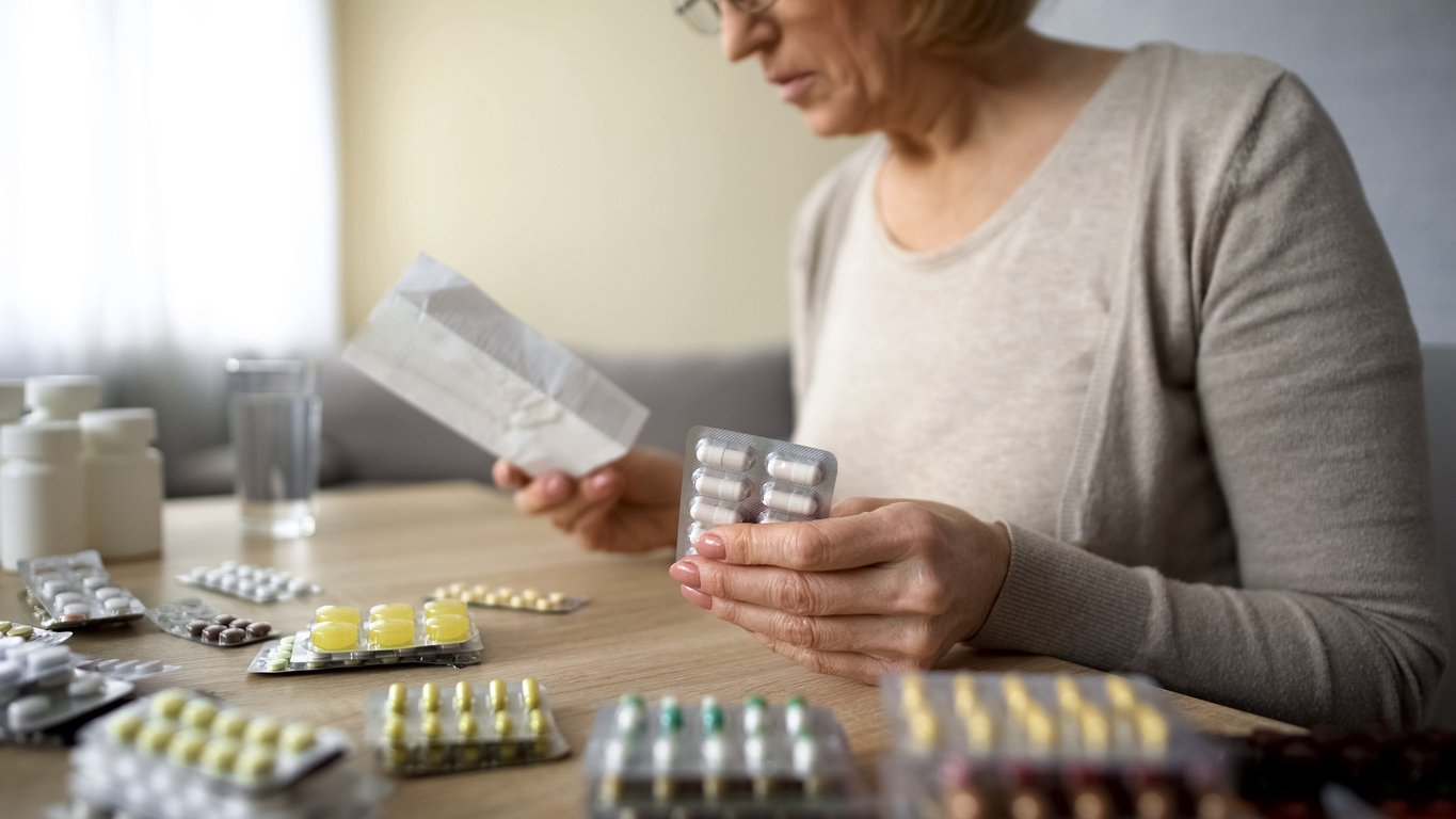 Home kitchen, elderly woman with medications, tabletop cluttered foreground, afternoon natural light, documentary style, people.