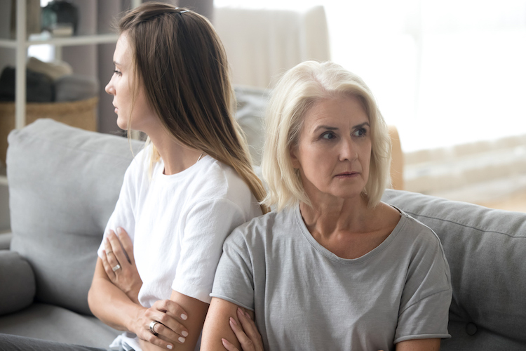 Living room, tense women side by side, crossed arms and averted gazes, daytime natural light, documentary style, two people.
