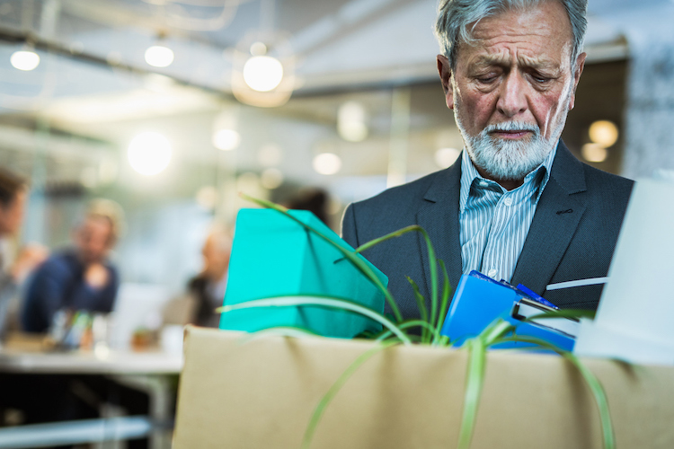 Office interior, elderly man with layoff box center frame, daytime soft light, documentary style, people present.