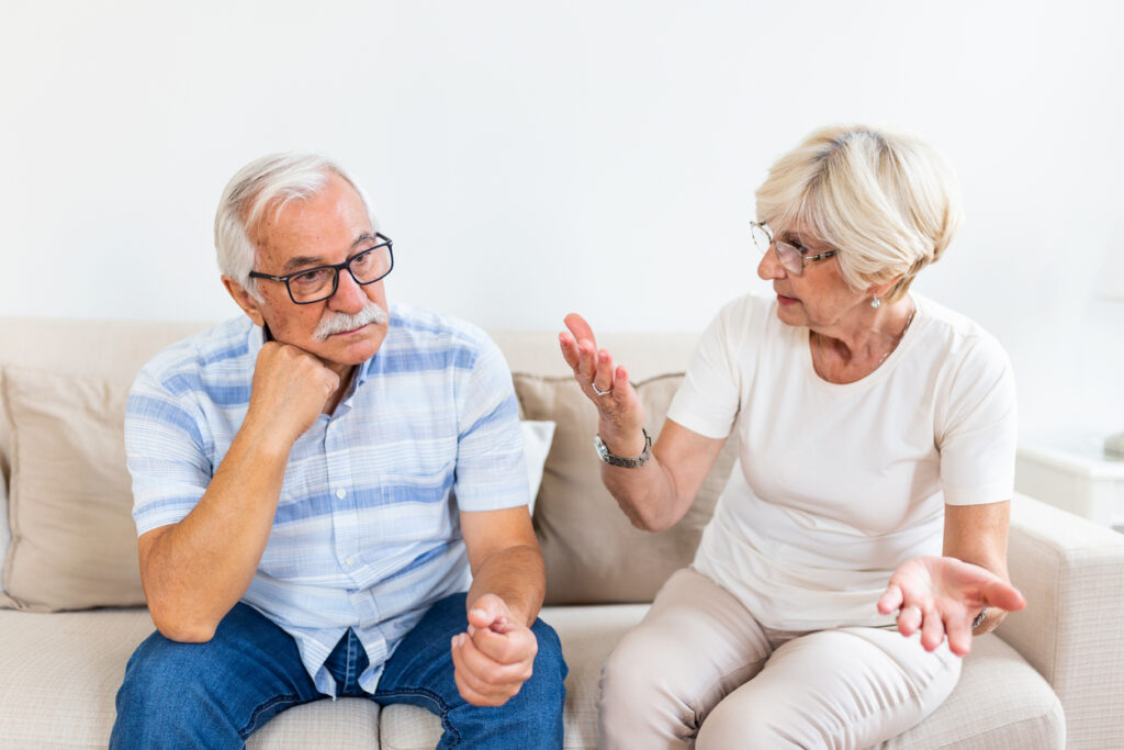 Living room couch, woman gesturing, man looking away, soft indoor daylight, documentary style, two older people.