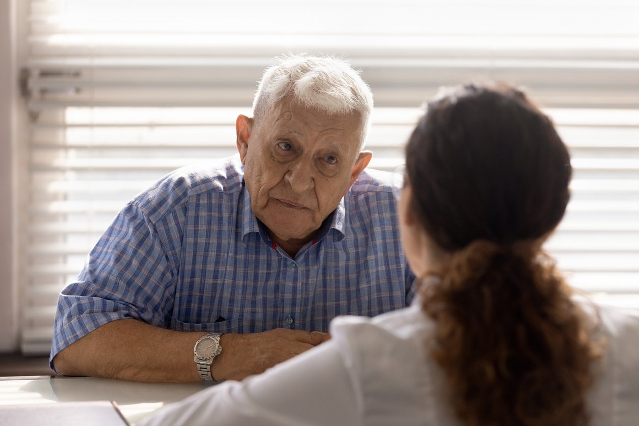 Bright room with window blinds, older man at table foreground, daylight, documentary style, doctor and patient present.