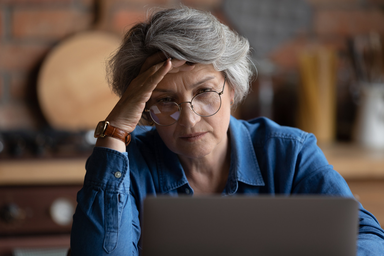 Kitchen interior, older woman with gray hair at table, laptop open, daylight, documentary style, single person.