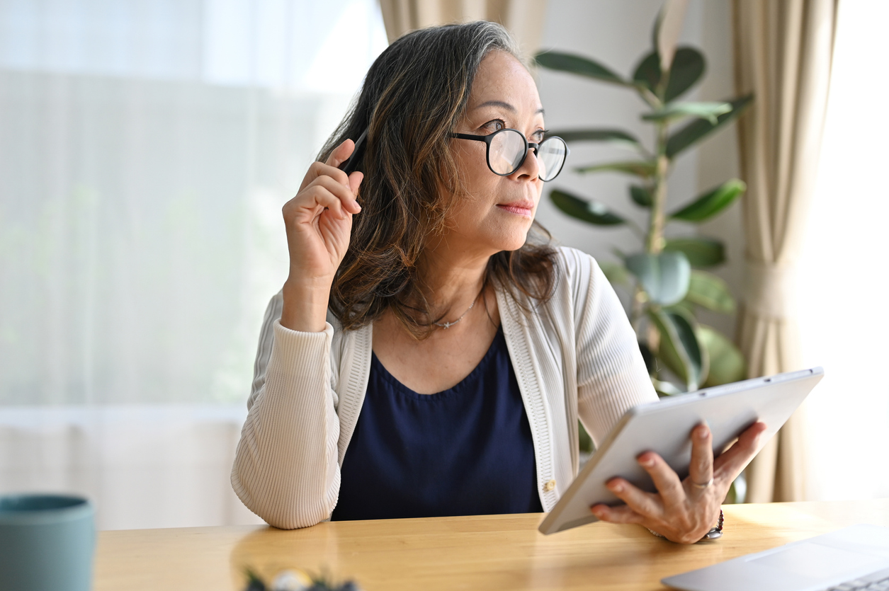 Dining room, older woman with glasses at table, half-profile with tablet, soft morning light, documentary style, one person.