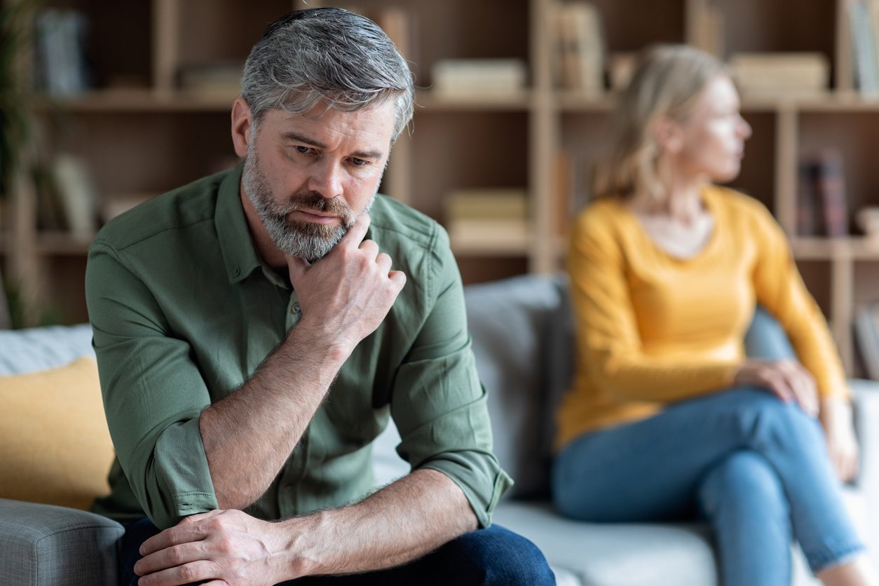 Living room, man with troubled face foreground, woman turned away beside him, daytime natural light, documentary style, two people.