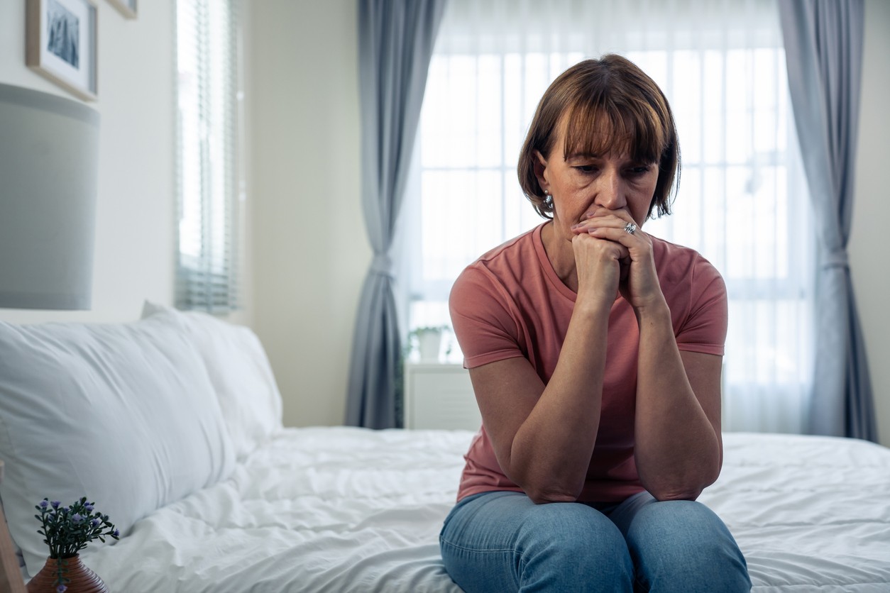 Bright bedroom, woman on bed edge, hands near mouth, morning light through window, documentary style, one person.