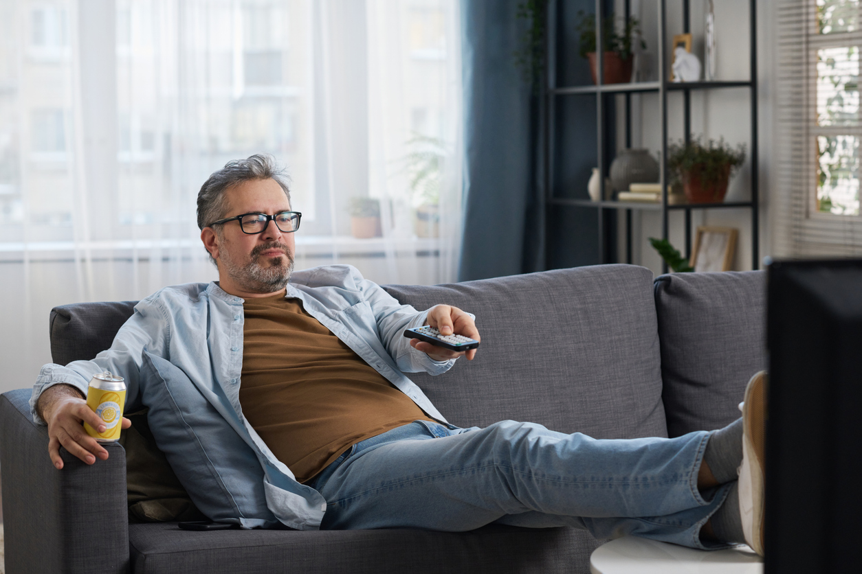 Bright living room, man with glasses on grey sofa, can and remote in hand, daytime light, documentary style, with people.