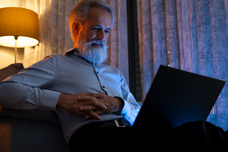 Living room, elderly man with white beard, on couch using laptop, nighttime lamp glow, documentary style, single person.
