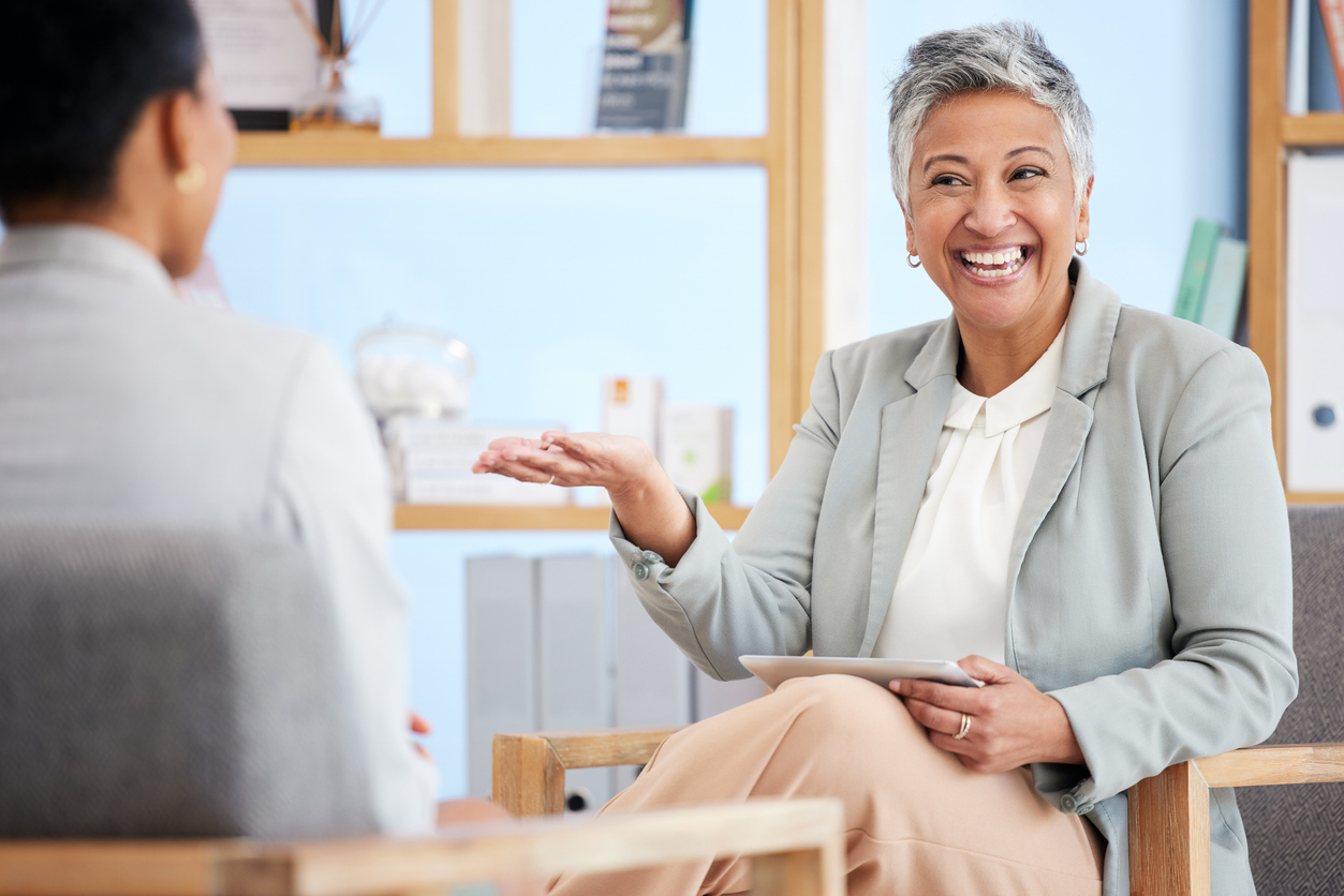 Office setting, gray-haired woman with tablet foreground, conversational pose, natural light, editorial workplace photo, two people.