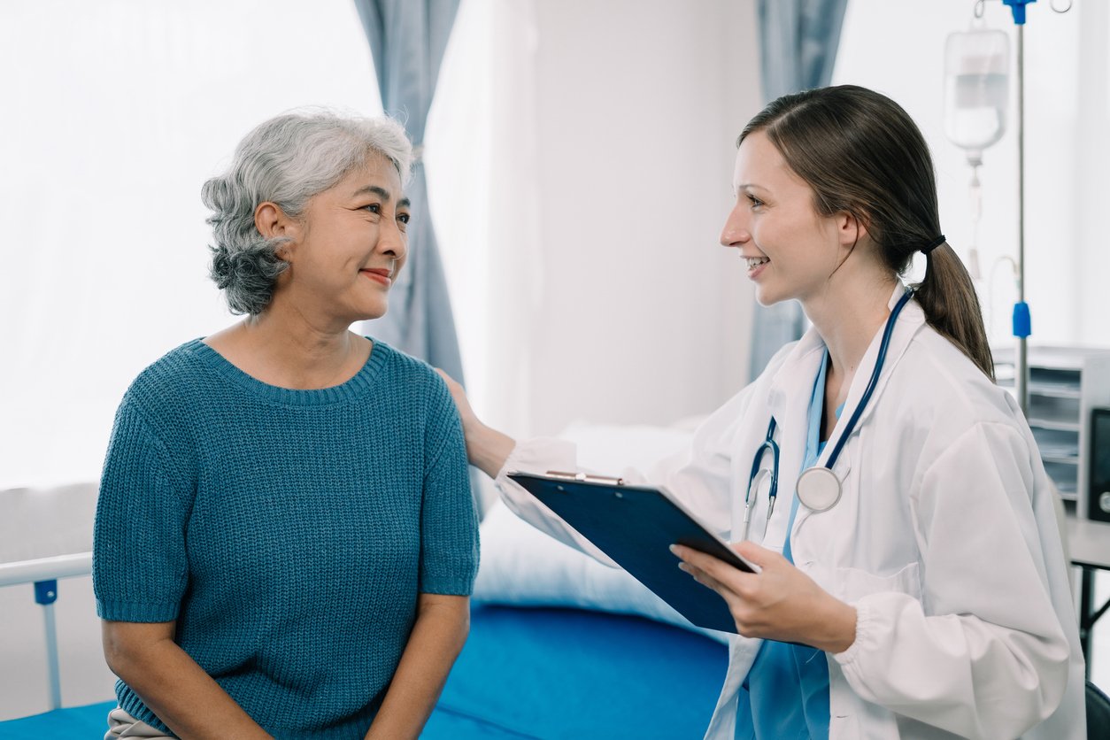 Bright medical office, doctor with clipboard and stethoscope facing older woman on exam bed, natural light, documentary style, people.