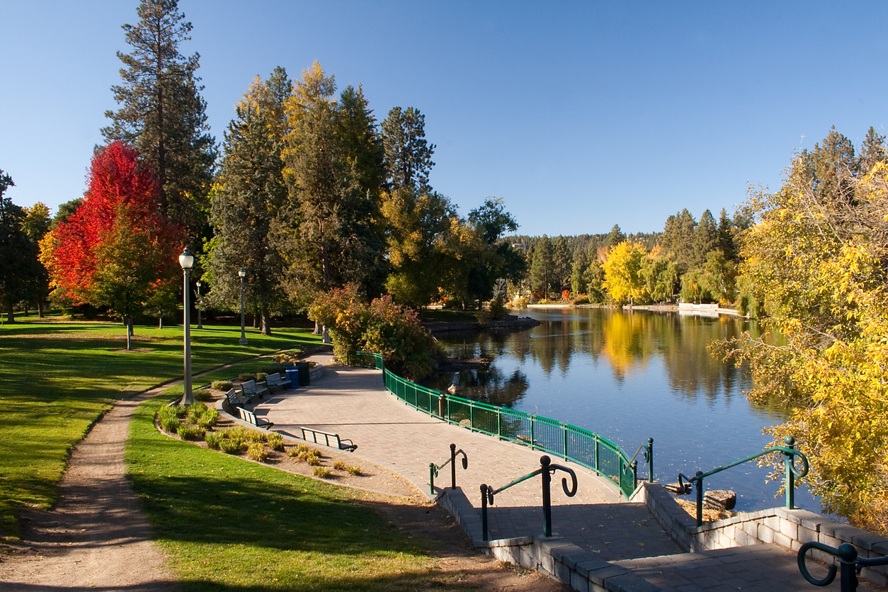 Park walkway, riverside focus, benches and trees frame scene, sunny afternoon with blue sky, editorial travel photo, no people.