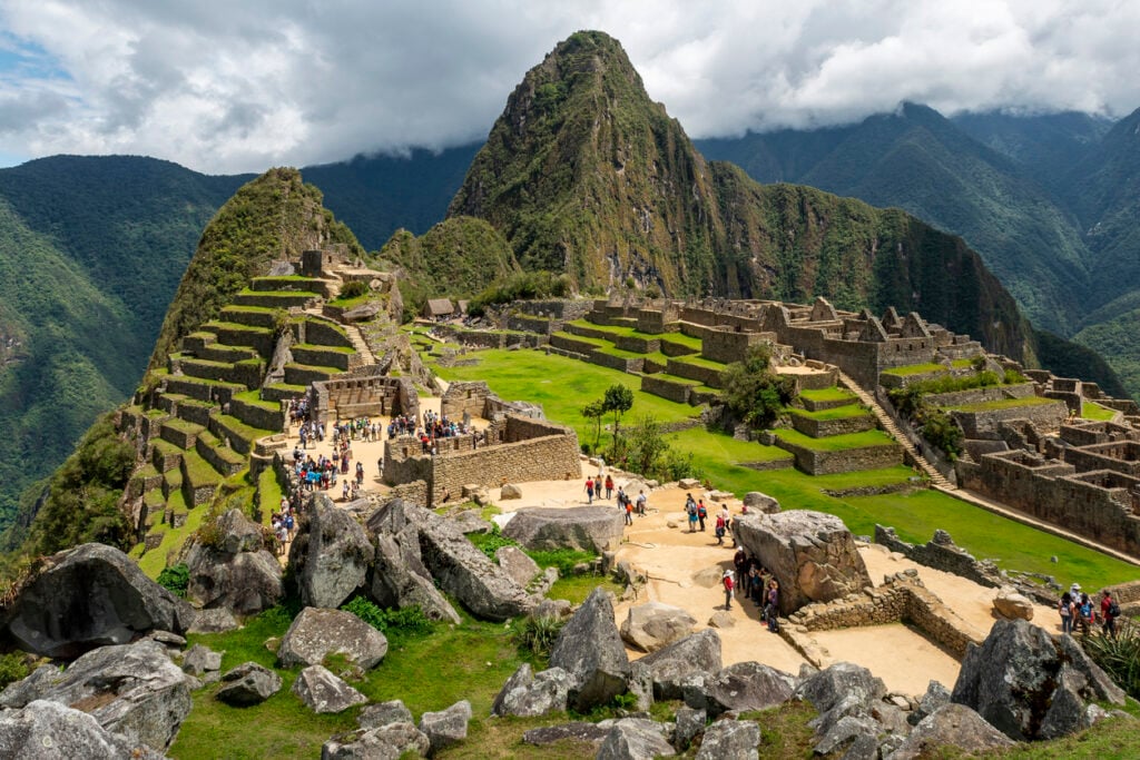 Machu Picchu, stone ruins center, terraced green slopes around, daytime clouds, editorial travel photo, tourists present.