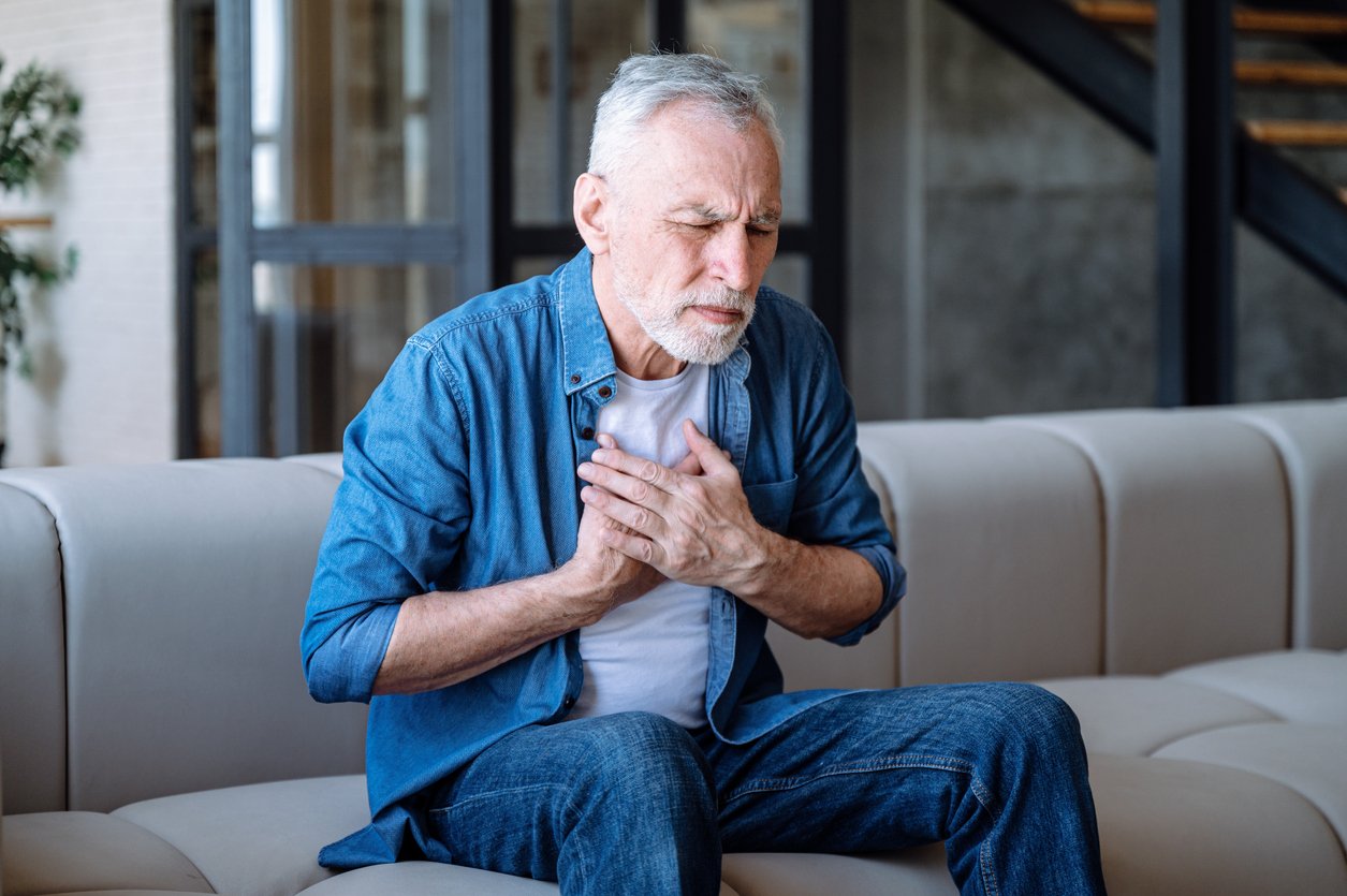 Living room, older man clutching chest, medium shot on couch, natural light, documentary style, one person.