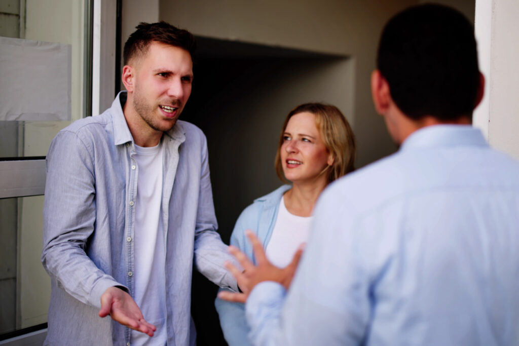 Doorway, three people talking, man gesturing in confusion, daytime light, documentary style, casual candid with people.