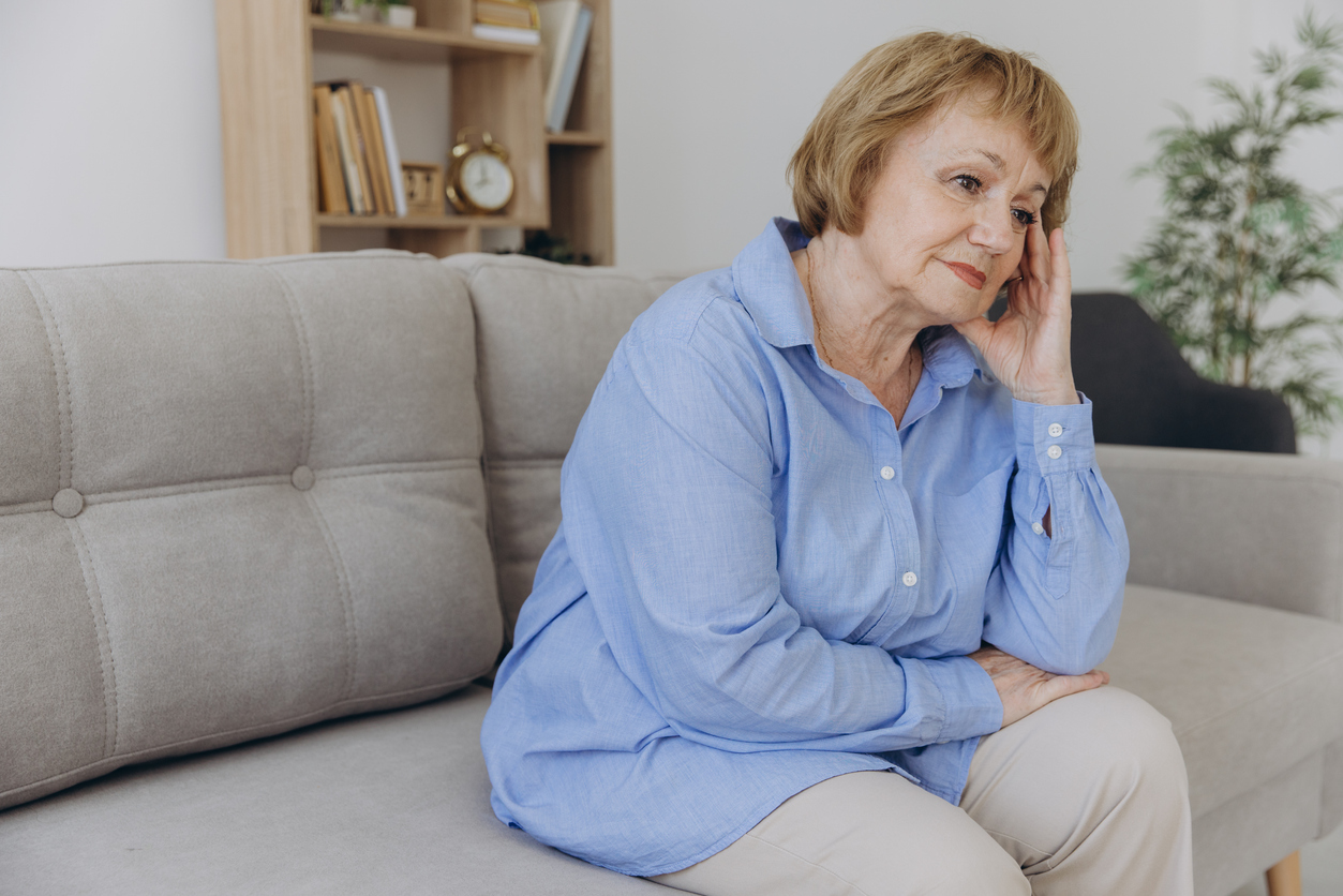 Living room, elderly woman in blue shirt on gray couch, thoughtful pose, daytime natural light, documentary style, people.