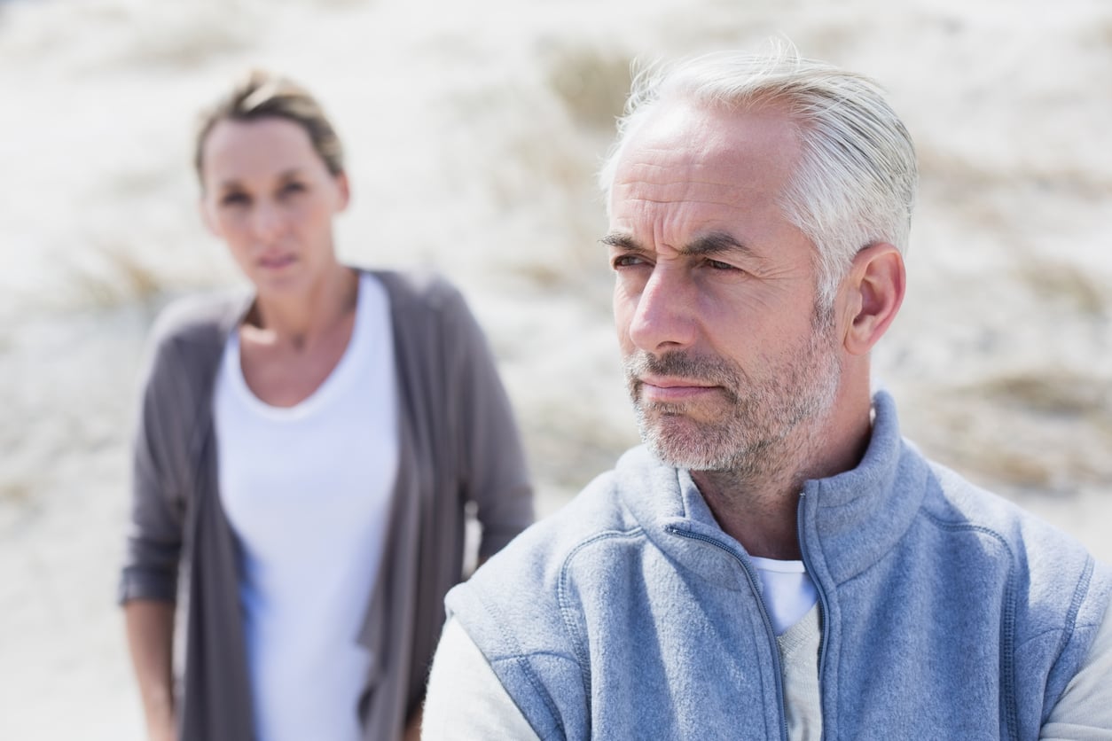 Sandy outdoor area, serious gray-bearded man foreground, woman blurred background, bright sunlight, editorial travel photo, two people.
