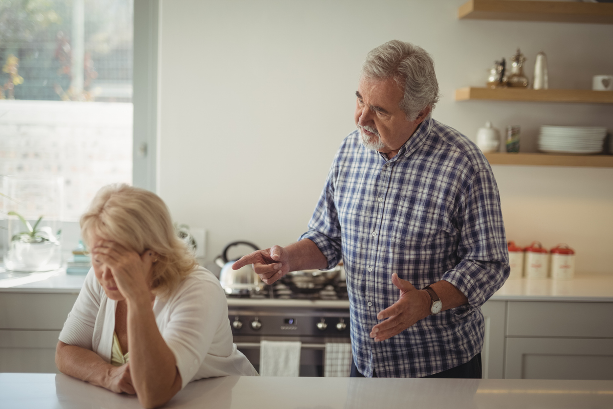 Kitchen interior, older man gesturing, woman head-in-hand at counter, midday natural light, documentary style, two people.