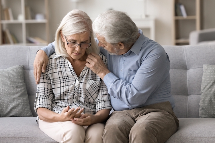 Living room, older couple on couch, man comforts woman, soft natural light, documentary style, two people.