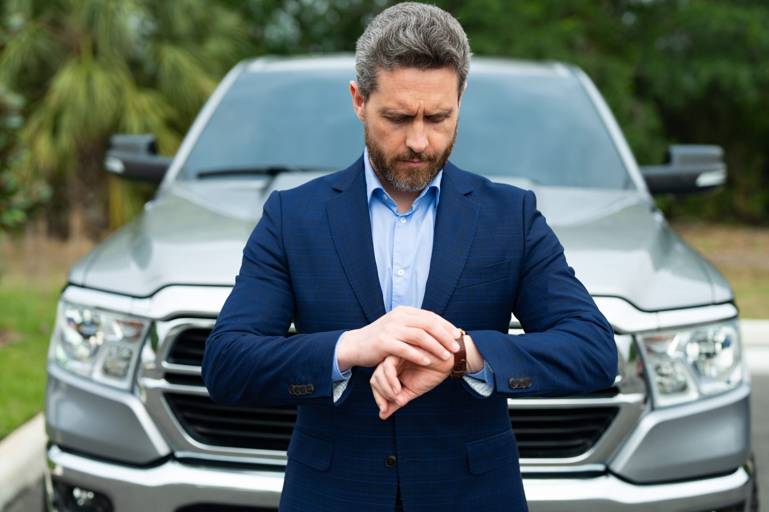 Outdoors with trees, blue-suited man checking wristwatch by silver pickup, foreground focus, daylight, editorial travel photo, people.
