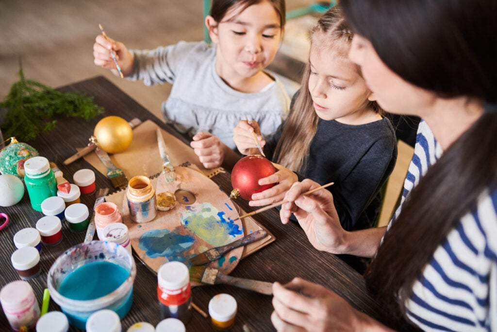 Festive room, Christmas ornaments painting, art supplies covering table, afternoon light, documentary style, three people.