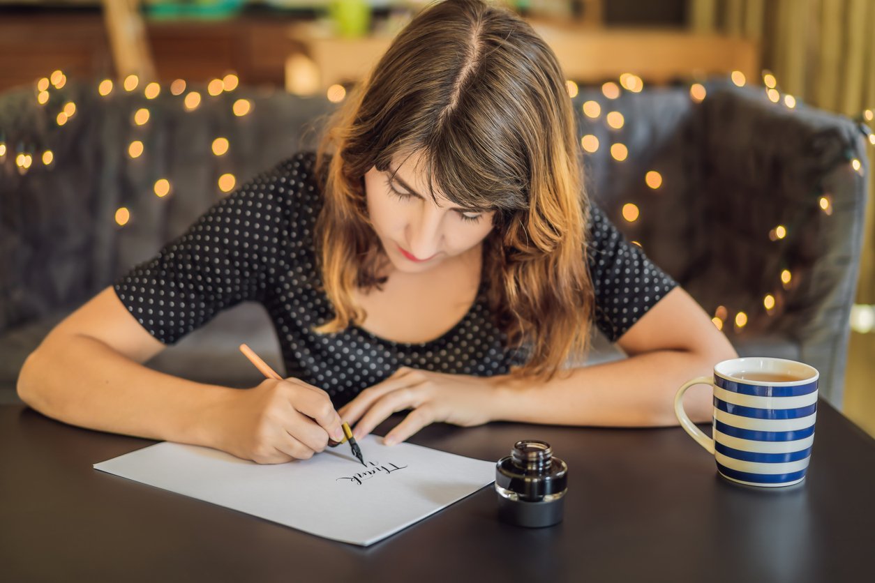 Cozy café, woman writing at table, cup and ink bottle foreground, warm dusk lights, editorial travel photo, one person.