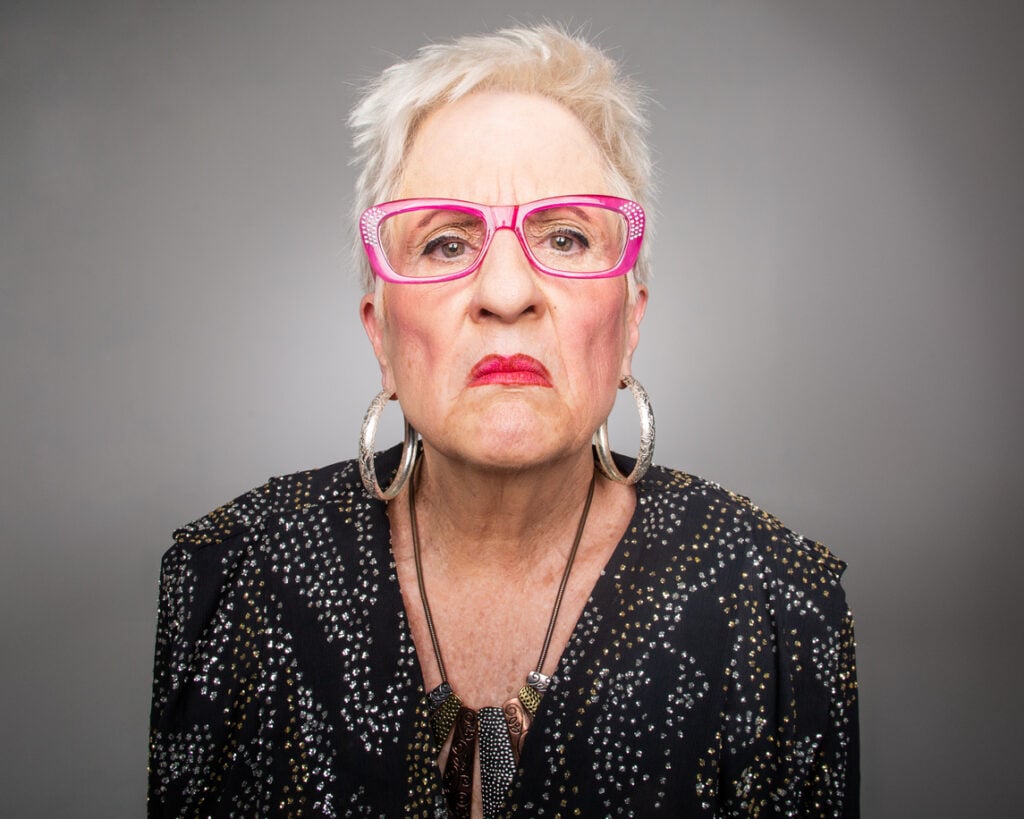 Studio portrait, pink glasses-wearing older woman, direct gaze, neutral lighting, editorial style, people.