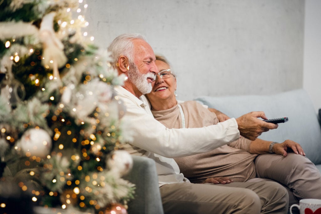 Living room, elderly couple on couch, Christmas tree foreground, cozy evening glow, documentary style, people.