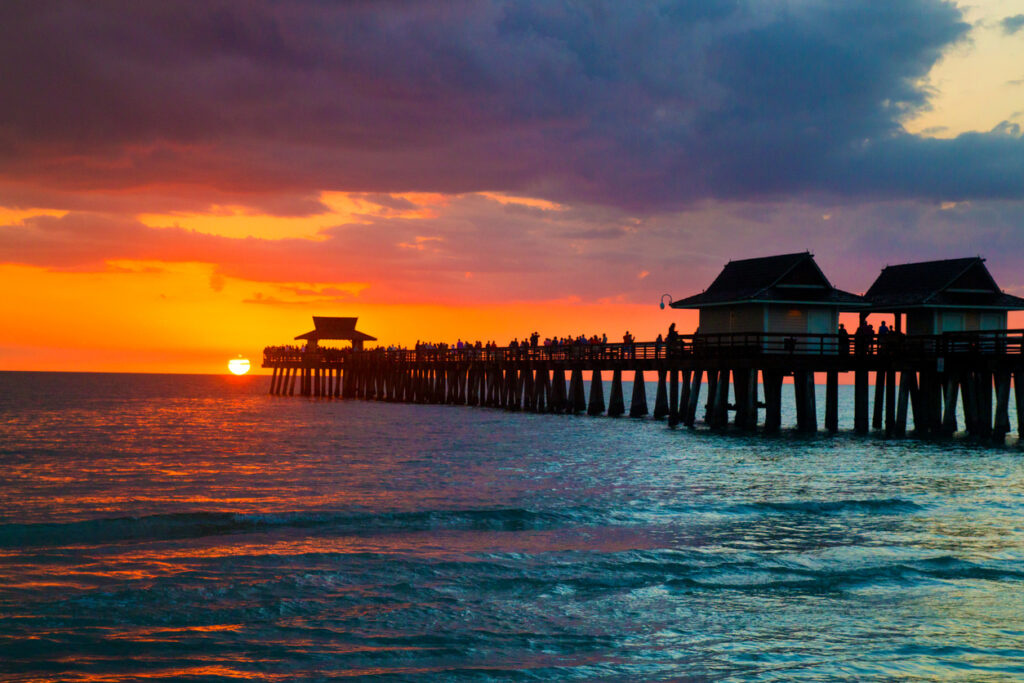 Ocean sunset, long pier, silhouetted people on walkway, golden hour glow, editorial travel photo, people visible.