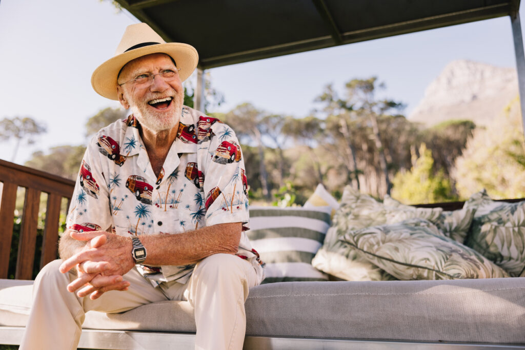 Outdoor terrace, older man in hat and patterned shirt foreground, wide shot, sunny afternoon, editorial travel photo, people.