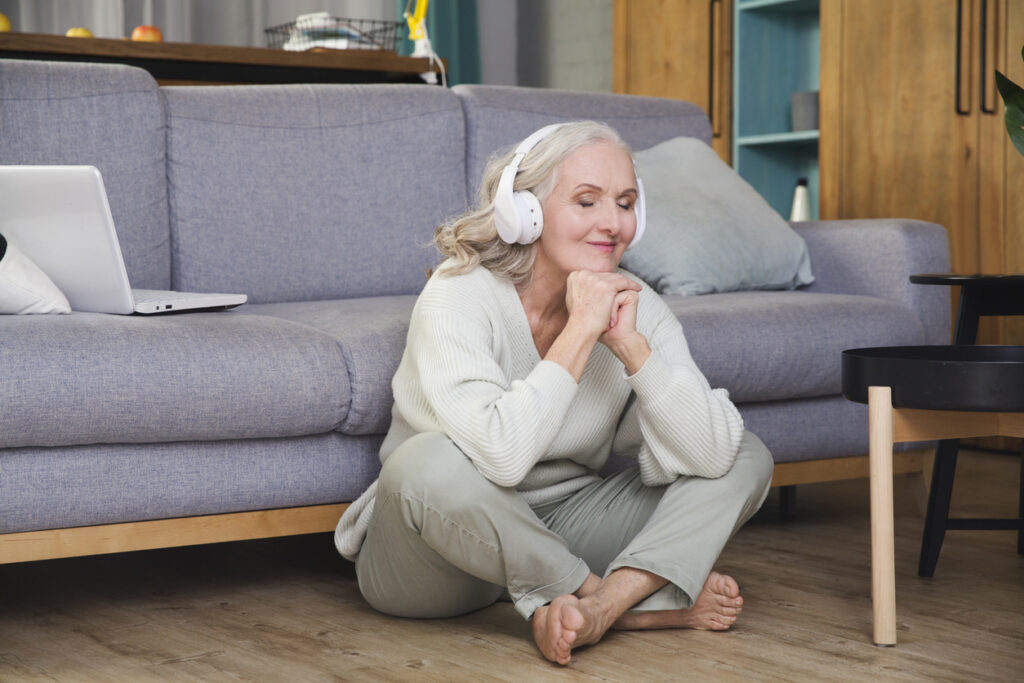 Cozy living room, gray-haired woman listening on floor, soft natural light, documentary style, relaxed mood, single person.