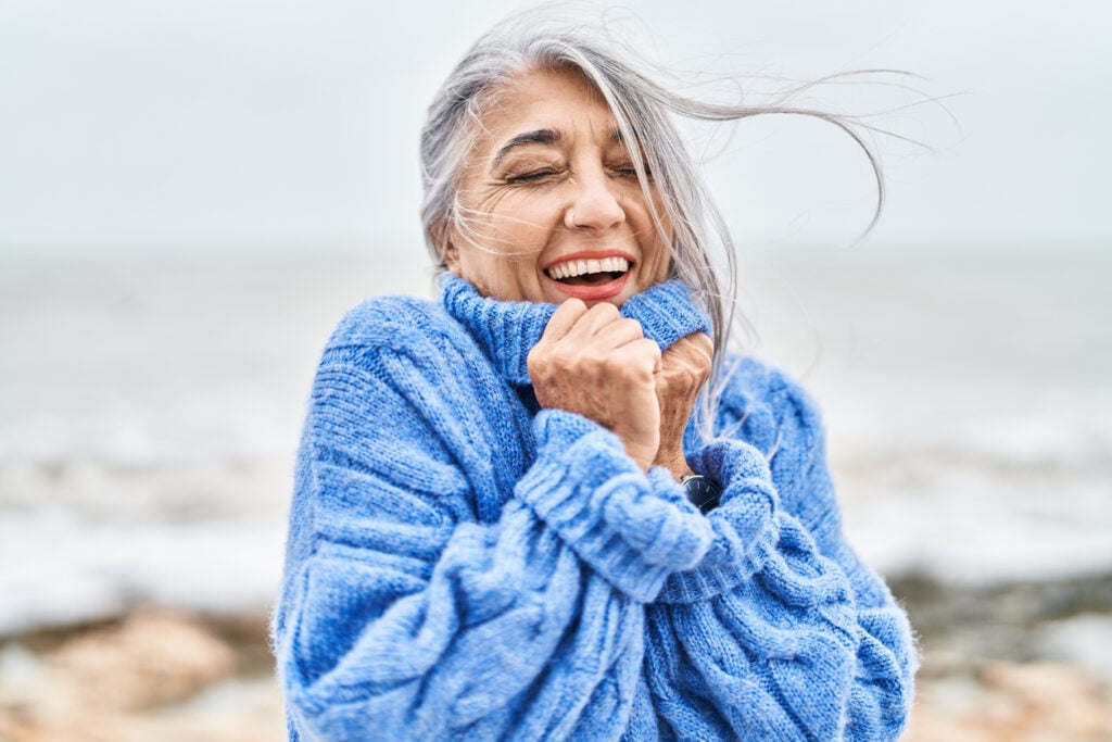 Blurry outdoor waterfront, joyful gray-haired woman pulls blue sweater collar, soft daylight, editorial travel photo, single person.