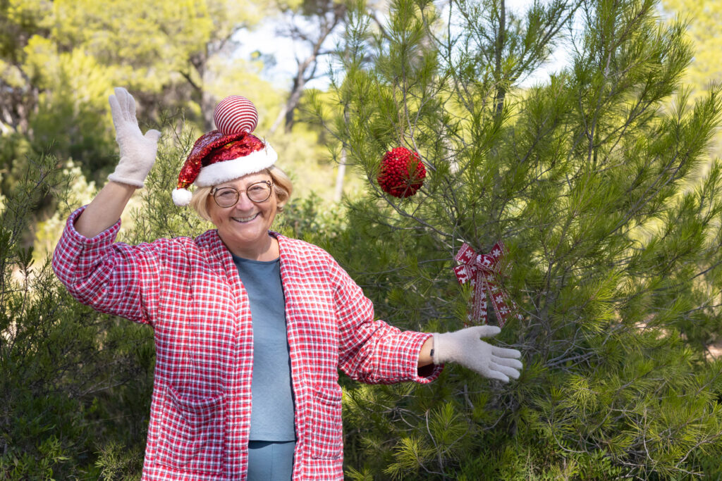 Outdoor setting, elderly woman in Santa hat waving, tree with red ornament beside her, daytime soft light, editorial travel photo, people.