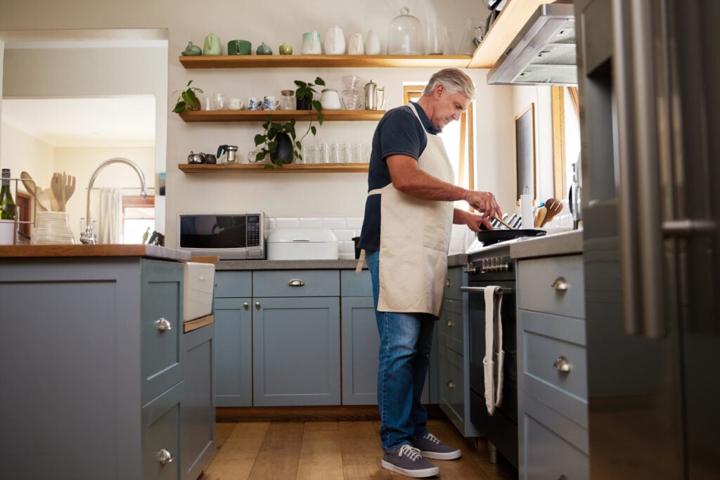 Modern kitchen, man at stove, blue cabinets and shelving, sunlit morning glow, editorial travel photo, single person.