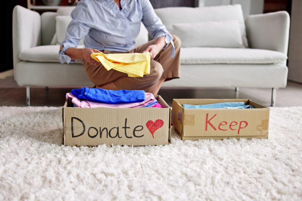 Living room, two donation boxes, clothes sorting foreground, soft daylight, documentary style, one person present.