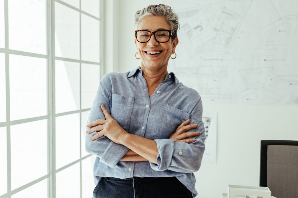 Bright office, older woman with glasses, arms crossed, daylight, documentary style, single person.