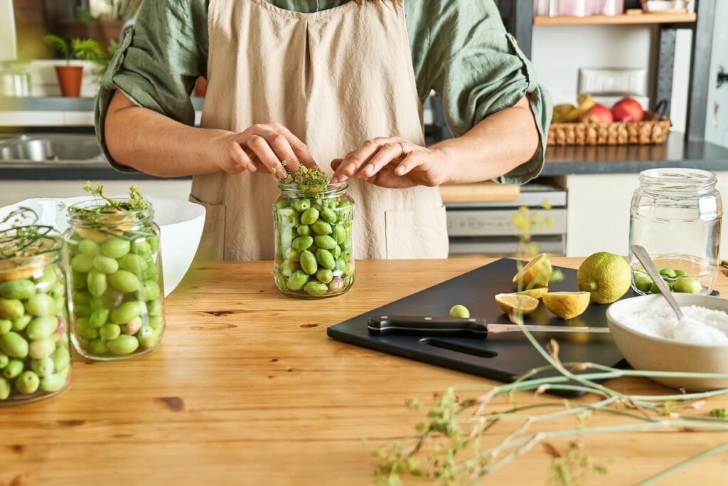 Kitchen counter, glass jars with green olives and herbs, overhead view, natural daylight, editorial travel photo, person present.
