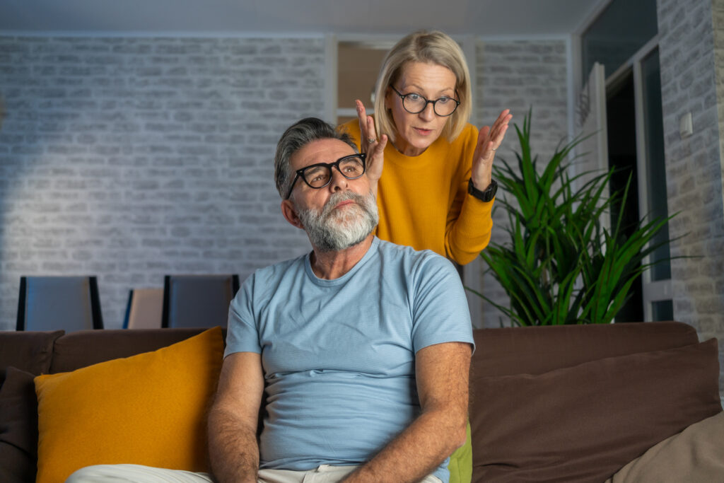 Modern living room, woman gesturing at man, both seated on couch, soft daylight, documentary style, two older people.